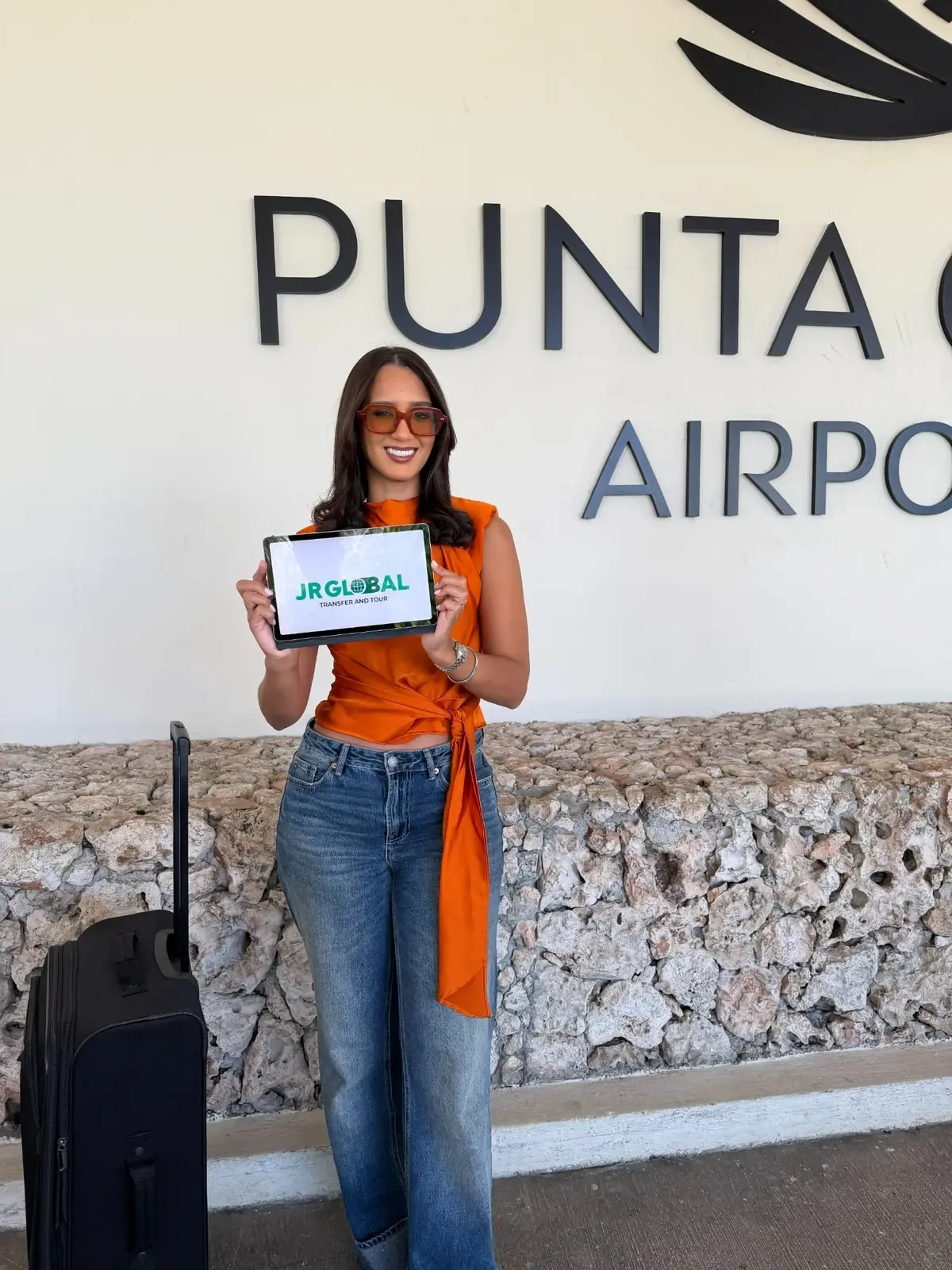 A woman with luggage and a JR Global Transfer and Tours tablet in front of the Punta Cana Airport sign.
